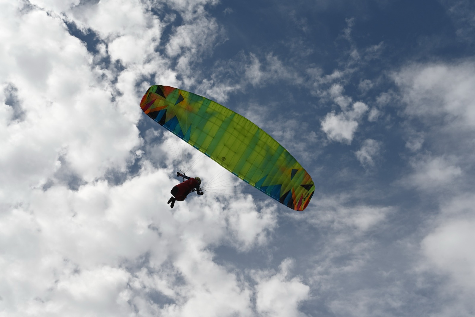 A paraglider under a green, orange, and blue wing against a cloudy sky